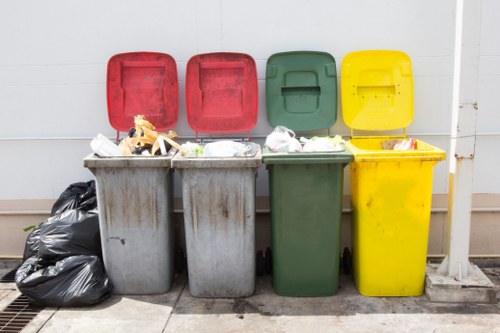 A row of four large wheelie bins situated outdoors against a light-colored wall, with their lids open or partially lifted. The first bin on the left is grey with a slightly weathered appearance, filled with mixed rubbish including plastic packaging and paper. Next to it, the second grey bin appears similar in size and condition, also containing waste. The third bin is green with a smoother finish, slightly less full and with the lid raised, revealing some rubbish inside. The fourth bin is bright yellow, plastic, with its lid open, exposing a small amount of waste within. To the left of the bins, there are two black garbage bags placed on the pavement, tied at the top. The setting appears to be an exterior space, possibly a back alley or side access area near a commercial property in East Ham, with a concrete ground and drainage grate visible in the foreground. This arrangement of waste receptacles reflects typical rubbish disposal for local businesses, with [COMPANY_NAME] providing collection services in the area.