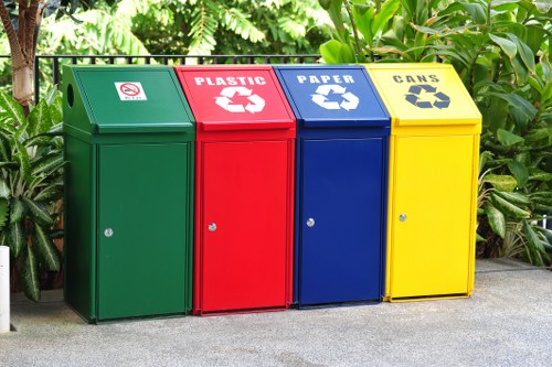 A set of four outdoor waste disposal bins positioned on a paved surface, each made of metal with a matte finish and different vibrant colours. From left to right, the first bin is green and designated for general waste, featuring a small icon of a crossed-out cigarette. Next is a red bin labeled for plastic recycling, displaying a white recycling symbol and the word 'PLASTIC' in bold uppercase letters. The third bin is blue, intended for paper waste, with a white recycling icon and the label 'PAPER.' The final bin is yellow, specified for cans, also featuring a white recycling symbol and the word 'CANS.' All four bins are upright, with small locking mechanisms on the front and slightly sloped lids. They are situated against a background of lush green foliage and tropical plants, suggesting an outdoor environment possibly in East Ham. The lighting indicates daytime with natural light illuminating the scene evenly. The presence of the bins aligns with rubbish collection services provided by Commercial Waste East Ham for local waste management, supporting environmentally conscious disposal practices near residential or commercial properties in the area.