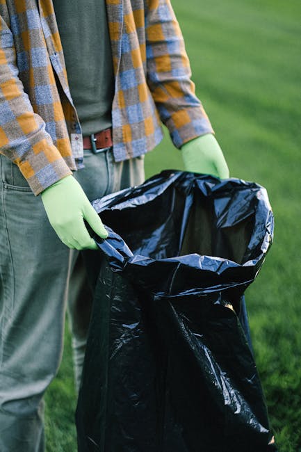 A group of three people, two adults and one child, are participating in a beach clean-up activity near the shoreline. The man, positioned on the right, is wearing a red t-shirt, light blue denim short