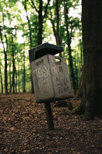 A green public rubbish bin with a rounded, dome-shaped lid partially transparent at the top, revealing discarded waste including paper wrappers and plastic items inside. The bin is mounted on a metal 