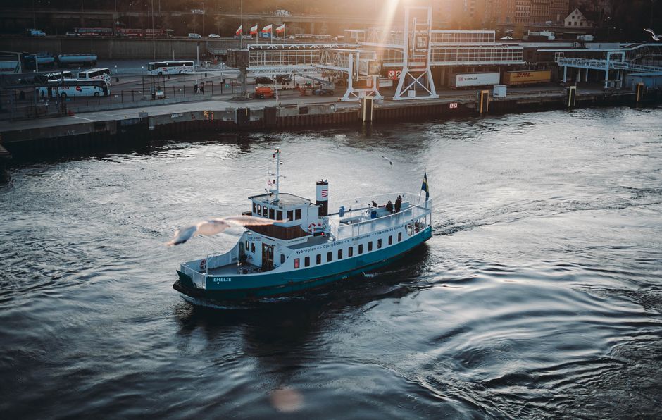 A small passenger ferry boat, predominantly painted in white and blue with a brown stripe along the side, is navigating through a river or harbor channel during late afternoon or early evening, as ind
